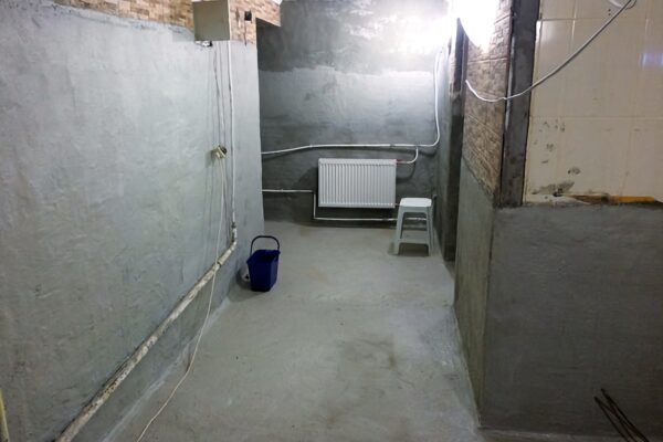 Підвал з відкритими трубами, радіатором і синім відром, нанесена гідроізоляція на стіни та підлогу.
