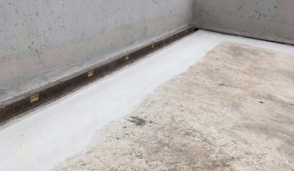 Бетонна поверхня з жолобом для стікання дощової води з ділянкою гідроізоляції, нанесеною вздовж стику підлоги зі стіною.